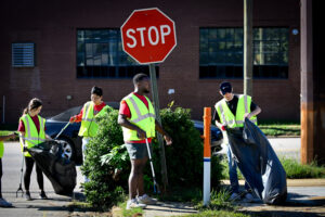 Four students in front of a stop sign with safety vests are holding trash bags and picking up trash.