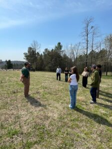 People form a semi-circle around a man in overalls in an open field.